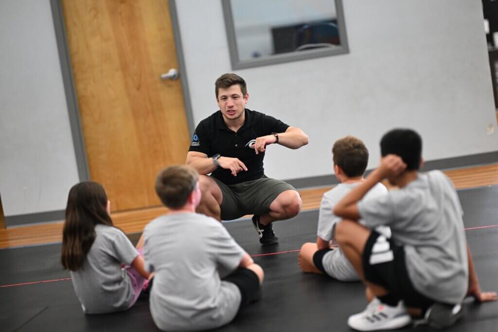 Coach crouching to talk with students seated on the gym floor